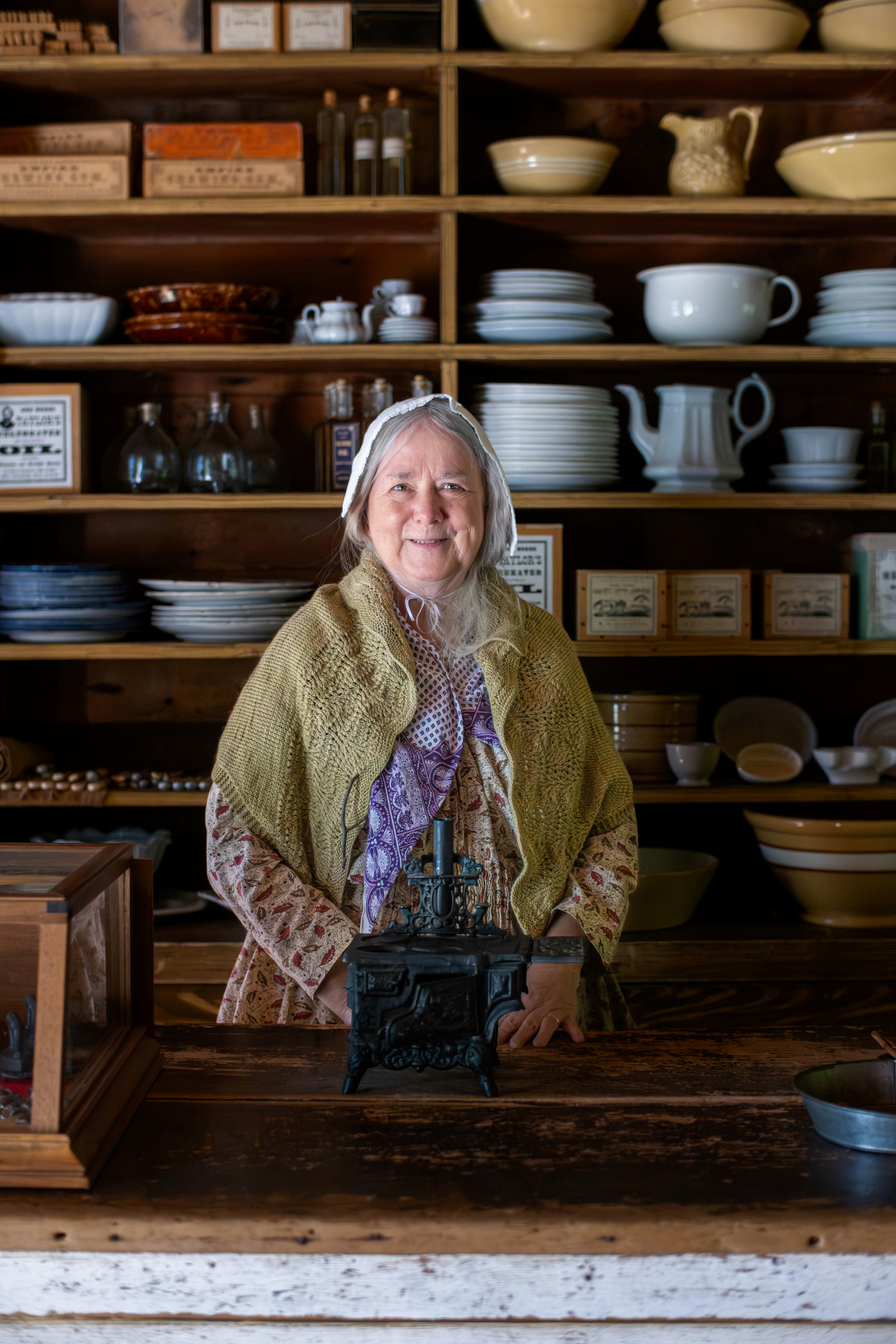 Woman in historic attire standing behind a counter with shelves of post and containers behind her.